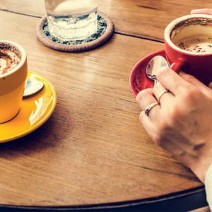 Kaffekopper på et bord, en rød og en gul kopp. Hender som holder kaffekoppene